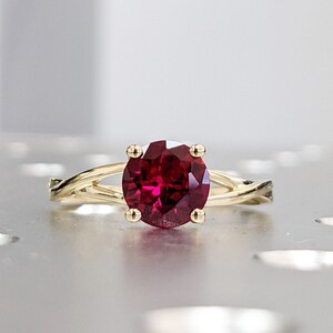 May include: A gold ring with a large, oval-cut, deep red gemstone. The band has a twisted design, with the gemstone held in place by four prongs. The ring is set against a light, neutral background.