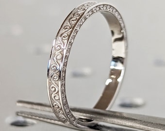 May include: A thin silver band with a delicate, intricate design around the edge.
