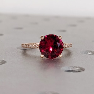 May include: A rose gold ring featuring a large, round, deep red gemstone set within a band of smaller, white gemstones.