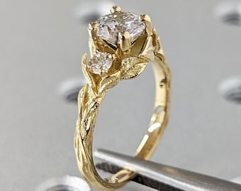 May include: A gold ring with a large, round, clear diamond in the center. The band is twisted and has a leaf design. There are two smaller diamonds on either side of the center diamond.