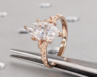 Könnte beinhalten: Ein Verlobungsring aus Roségold mit einem großen, klaren Diamanten in Marquise-Form. Der Ring hat ein zartes Blumenmuster auf der Band.