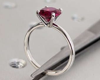May include: A silver ring with a red gemstone set in a prong setting. The ring is held by a pair of tweezers.
