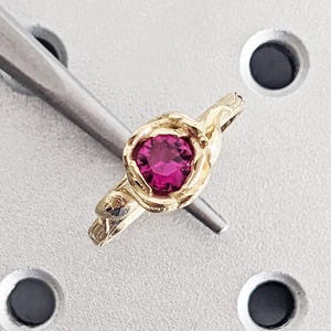 Pode incluir: Um anel de ouro com uma gema rosa vibrante e redonda. O anel tem uma banda e uma cravação texturizadas, com um design detalhado e orgânico. O anel está sobre uma superfície cinza claro.