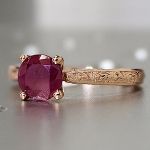 May include: A rose gold ring featuring a deep red ruby gemstone. The band is engraved with a delicate floral design.