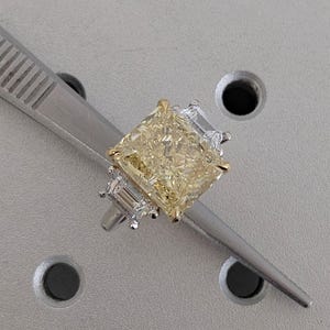 May include: A yellow radiant-cut diamond ring with a gold band and two smaller, clear side stones. The ring is held by a silver tweezer against a grey background. The diamond has a square shape with a brilliant cut.