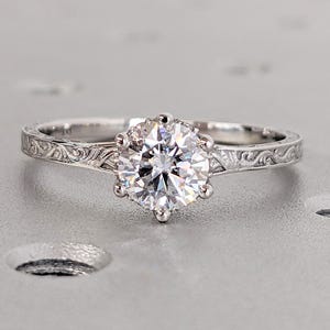 Pu&ograve; includere: Un anello di fidanzamento in argento con una grande gemma rotonda e trasparente. L'anello ha un'incastonatura a sei punte e una fascia con un motivo floreale inciso. L'anello &egrave; esposto su una superficie grigio chiaro.