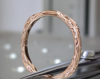 Puede incluir: Un anillo de banda de oro rosa con un diseño detallado e intrincado. El anillo está sujeto por unas pinzas.