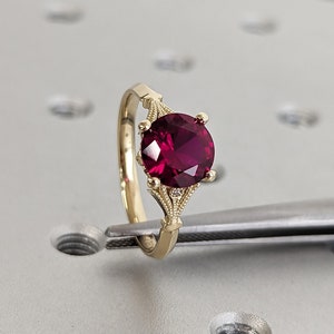 Puede incluir: Un anillo de oro con una gran piedra de rubí redonda y de color rojo intenso engastada en un diseño delicado y ornamentado. El anillo está sujeto por unas pinzas sobre una superficie gris.