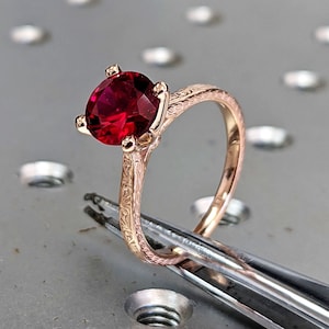 May include: A rose gold engagement ring with a red gemstone set in a filigree band. The ring is held by tweezers on a metal surface.