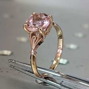 May include: A delicate gold ring with a large, oval-shaped pink gemstone set in a detailed, ornate design.