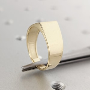 May include: A gold signet ring with a flat, rectangular top. The ring is being held by a pair of tweezers.