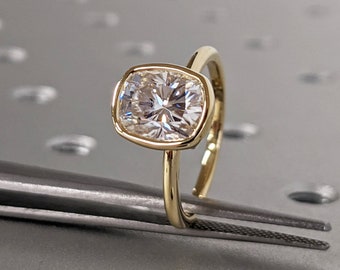 Könnte beinhalten: Ein goldener Ring mit einem großen, quadratisch geschliffenen, klaren Edelstein. Der Edelstein ist in einer Lünettenfassung gefasst. Der Ring liegt auf einer grauen Oberfläche, gehalten von einer Pinzette.