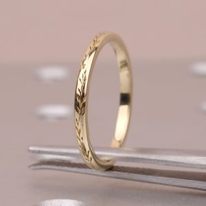 May include: A delicate gold ring with an engraved leaf pattern. The ring is held by a pair of silver tweezers, showcasing its intricate design. The ring's slender band and polished finish highlight its elegant simplicity, perfect for everyday wear.