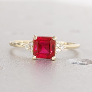 Pode incluir: Um anel de ouro com uma pedra vermelha quadrada e pedras transparentes menores na banda.