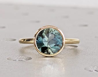 May include: A gold ring featuring a round, faceted teal gemstone. The gemstone is set in a bezel setting, and the band is a simple, polished gold. The ring is displayed on a light gray surface.