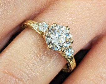 May include: A gold engagement ring with a large round diamond and two smaller diamonds on the band. The ring is on a finger.