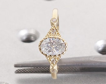 May include: A gold ring with an oval-shaped diamond set in a delicate, detailed setting. The diamond is surrounded by smaller diamonds, and the band is adorned with a delicate pattern.