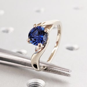 Könnte beinhalten: Ein silberner Ring mit einem blauen Saphir-Edelstein. Der Ring hat ein verdrehtes Band und einen runden, facettierten Stein.