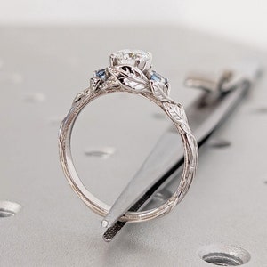 May include: A silver ring with a large, round, clear gemstone surrounded by smaller blue gemstones. The ring is set in a delicate, leafy design.