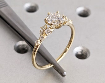 Op de afbeelding: Een delicate gouden verlovingsring met een ronde middelste steen en kleinere stenen die langs de band zijn gezet.