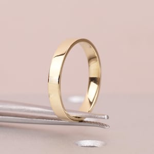 May include: A polished gold band ring, held by tweezers, against a soft pink background. The ring has a simple, classic design with a smooth, reflective surface. The ring's width is approximately 0.2 cm.