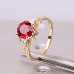 May include: A gold ring featuring a large, round, faceted red gemstone as the center stone, flanked by two smaller, clear, round, faceted stones. The band is crafted with a textured, organic leaf-like design that wraps around the finger.