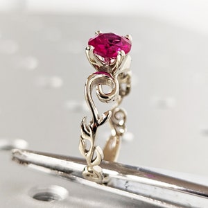 Op de afbeelding: Een gouden ring met een robijn-edelsteen gezet in een bloemmotief. De ring ligt op een wit oppervlak.