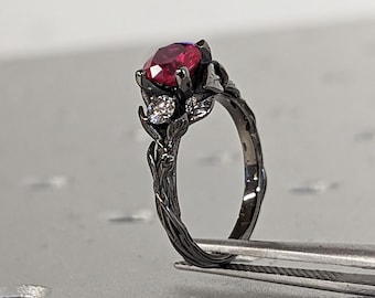 May include: A black ring with a red gemstone and a leaf design. The ring is held by a pair of tweezers.