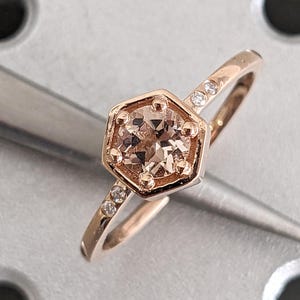 May include: A rose gold ring featuring a central round champagne-colored gemstone set within a hexagonal bezel. The band is accented with two small clear round stones on each side of the central setting, totaling four accent stones.