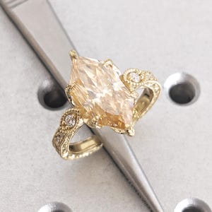Könnte beinhalten: Ein goldener Ring mit einem großen, marquiseförmigen, hellgelben Edelstein als Mittelstück. Der Ring weist aufwendige Details mit kleineren, runden, klaren Edelsteinen und kunstvollen Filigranarbeiten am Band auf.
