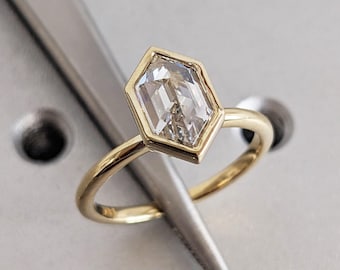 May include: A gold ring featuring a hexagonal-cut, clear gemstone. The gemstone is set in a gold bezel, and the band is a simple, polished gold. The ring is shown against a light-colored, textured background, with a silver tool in the frame.