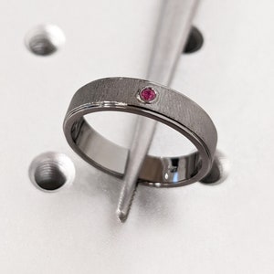 Puede incluir: Un anillo de plata con acabado cepillado y una pequeña piedra preciosa roja engastada en el centro.