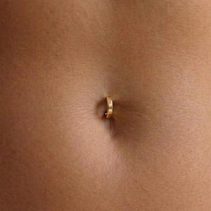 May include: A small, polished gold-toned circular hoop belly button ring is inserted into the navel. The jewelry features a simple, continuous band design with a smooth, reflective metallic finish, positioned centrally within the navel area.