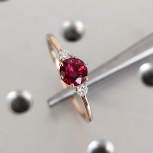 Può includere: Un anello in oro rosa con una grande gemma rosso scuro rotonda, affiancata da due gemme chiare più piccole. L'anello è tenuto da uno strumento d'argento su uno sfondo grigio chiaro. La fascia dell'anello è sottile e delicata.