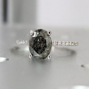 May include: A silver ring with a large, oval-shaped, black diamond. The diamond has a unique, faceted design and is surrounded by a band of smaller diamonds.