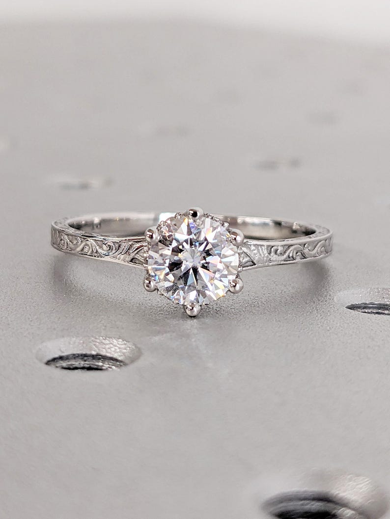 Pu&ograve; includere: Un anello di fidanzamento in argento con una grande gemma rotonda e trasparente. L'anello ha un'incastonatura a sei punte e una fascia con un motivo floreale inciso. L'anello &egrave; esposto su una superficie grigio chiaro.