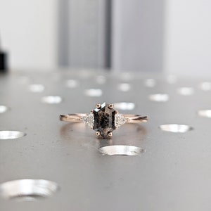 May include: A rose gold engagement ring with a gray octagon-shaped diamond and two smaller triangular diamonds on either side.