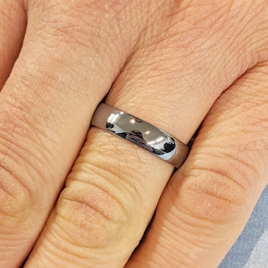 May include: A shiny, silver-colored ring with a smooth, rounded surface. The ring is worn on a finger, reflecting the surrounding environment. The ring's design is simple and elegant, suitable for everyday wear.