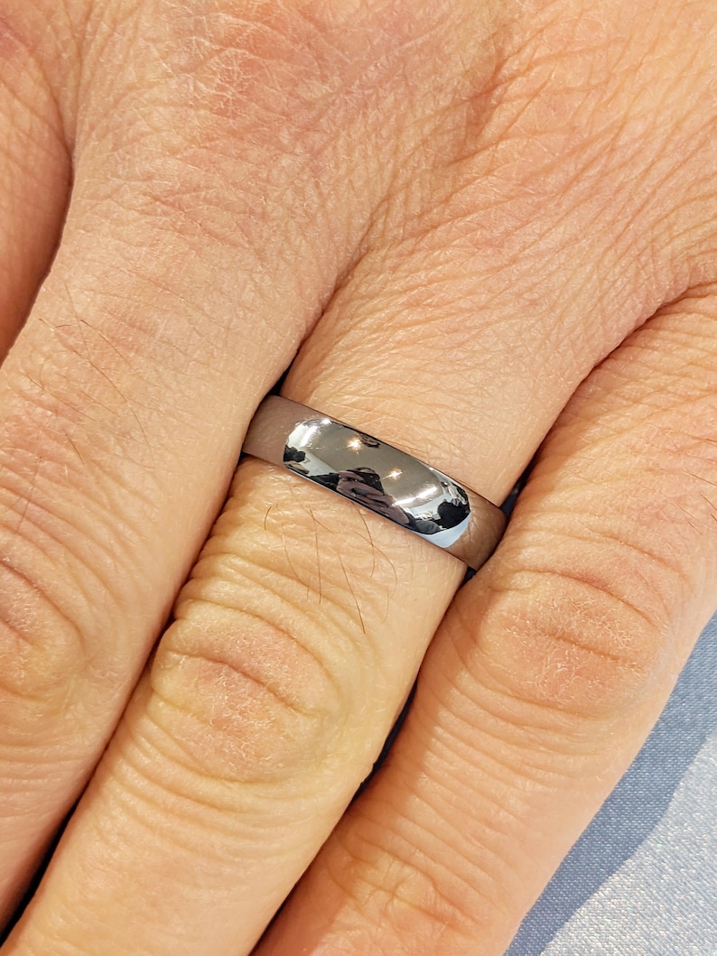 May include: A shiny, silver-colored ring with a smooth, rounded surface. The ring is worn on a finger, reflecting the surrounding environment. The ring's design is simple and elegant, suitable for everyday wear.