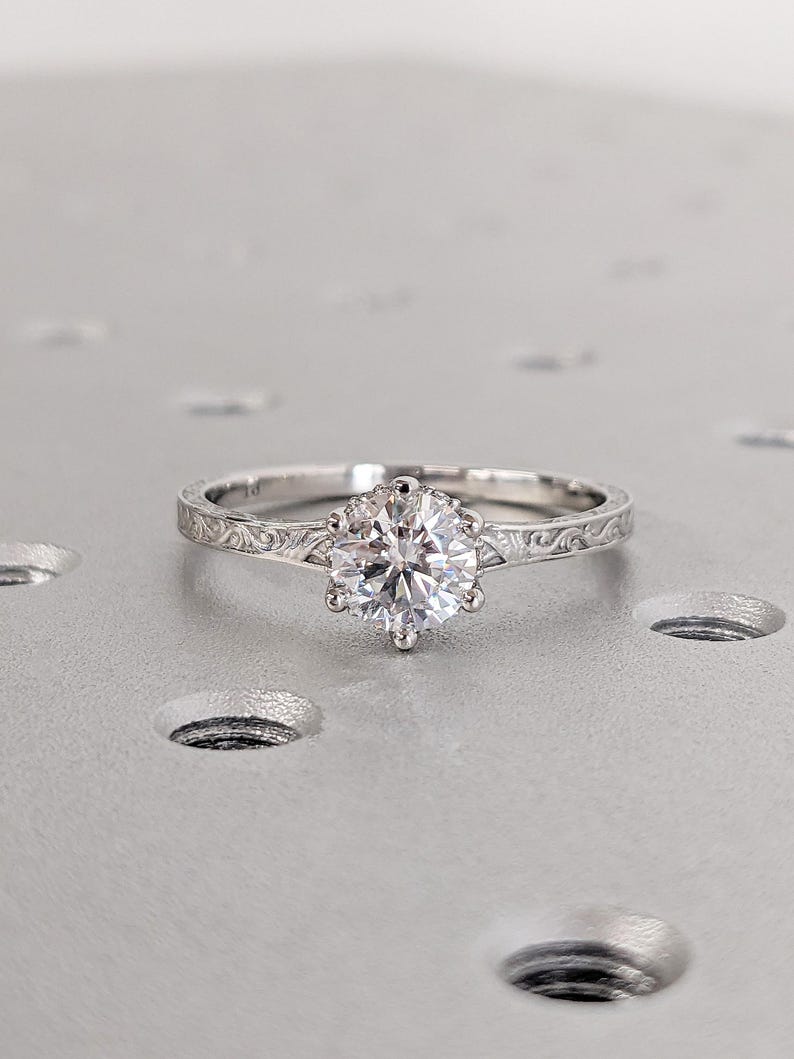 Pu&ograve; includere: Un anello di fidanzamento in argento con una grande gemma rotonda e trasparente. L'anello ha un'incastonatura a sei punte e una fascia incisa dettagliata. L'anello &egrave; appoggiato su una superficie grigio chiaro.