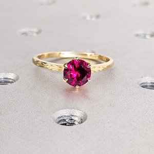 Puede incluir: Un anillo de oro con una gran gema redonda de color rosa intenso. La banda tiene un diseño delicado y grabado. El anillo se encuentra sobre una superficie gris claro con hendiduras circulares.