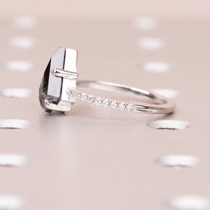 Puede incluir: Un anillo de plata con una gema oscura de forma irregular. La banda est&aacute; adornada con peque&ntilde;as piedras brillantes, a&ntilde;adiendo un toque de elegancia. El anillo se encuentra sobre un fondo suave y neutro.