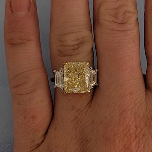 May include: A ring featuring a large, radiant-cut yellow diamond, with two smaller rectangular white diamonds on either side. The central yellow diamond is the focal point. The setting appears to be a combination of gold and platinum.