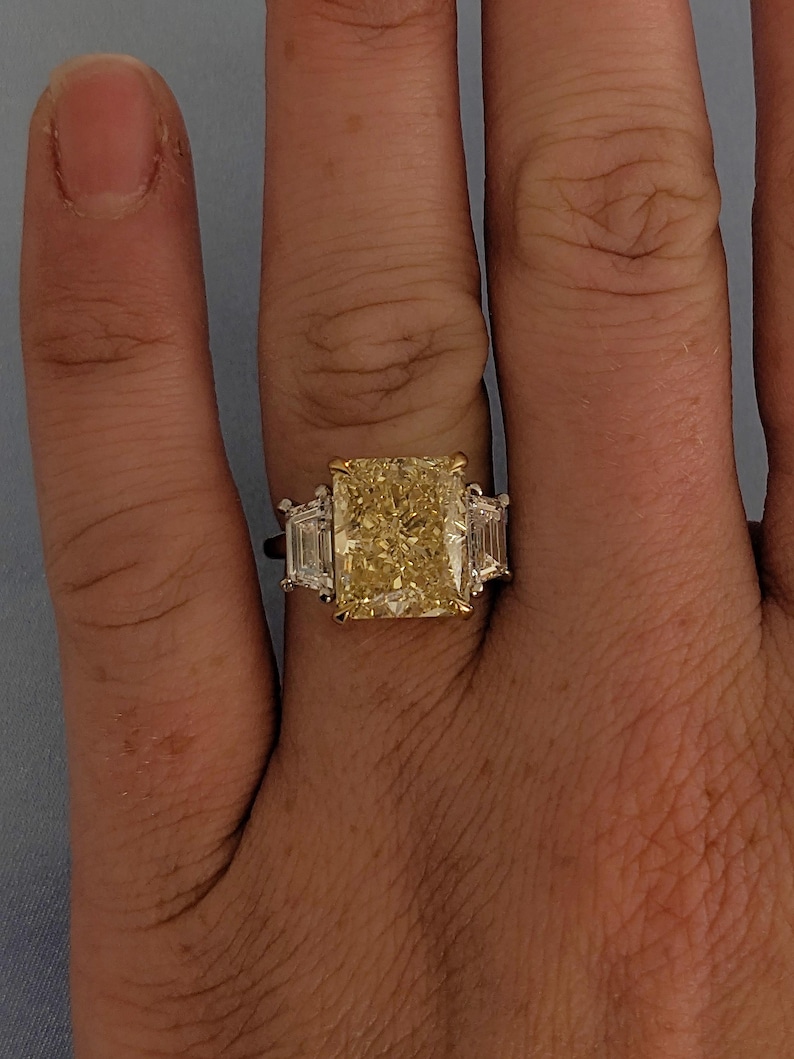 May include: A ring featuring a large, radiant-cut yellow diamond, with two smaller rectangular white diamonds on either side. The central yellow diamond is the focal point. The setting appears to be a combination of gold and platinum.