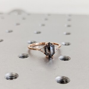 May include: A rose gold ring with a dark grey octagon-shaped gemstone and two smaller white triangular gemstones on either side.