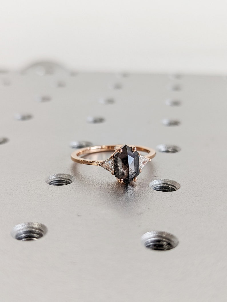 May include: A rose gold ring with a dark grey octagon-shaped gemstone and two smaller white triangular gemstones on either side.