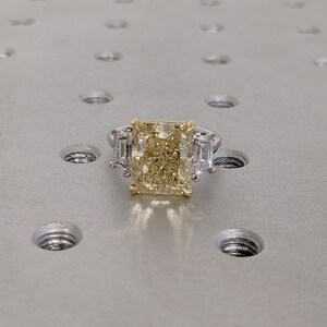 May include: A radiant-cut yellow diamond ring with two baguette-cut side stones. The ring features a platinum band and a gold setting for the center stone. The ring is set on a textured, silver surface.