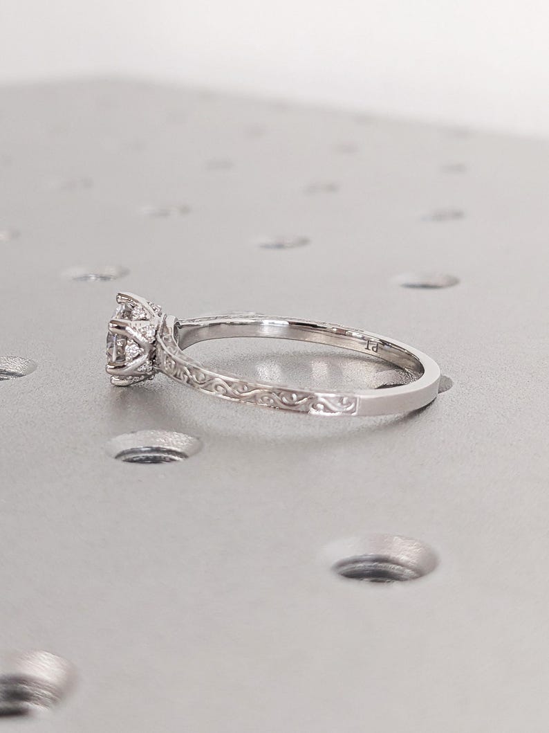 Pu&ograve; includere: Un anello di fidanzamento in argento con una gemma rotonda e trasparente e dettagli di filigrana intricati. La fascia presenta delicate volute. L'anello &egrave; posto su uno sfondo grigio chiaro.