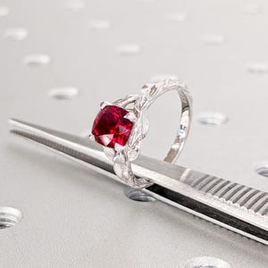 Puede incluir: Un anillo de plata con una piedra preciosa roja engastada en un diseño floral. El anillo está apoyado sobre una superficie plateada.