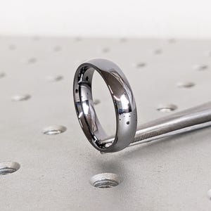 May include: A polished, silver-colored ring with a smooth, rounded design. The ring is held by a metal tool, showcasing its reflective surface. The ring's simple design suggests it could be a wedding band or a fashion accessory.